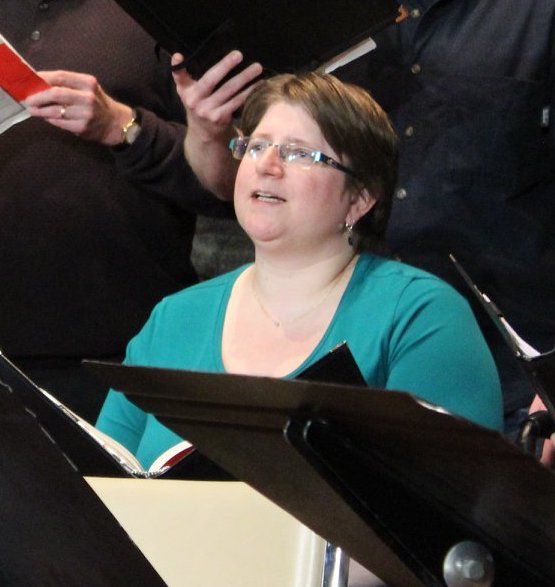 Diane chante, assise sur une scène, entourée de chanteurs debout. Elle porte un haut vert et tient un classeur noir contenant sa partition musicale.